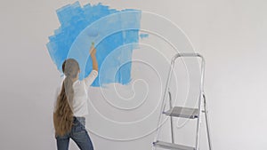 Young girl paints the wallpaper with a roller in the room with blue paint
