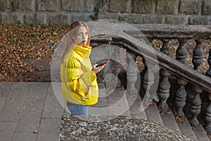 A young girl with phone, in yellow jacket, on the stairs of an old building. Autumn day.