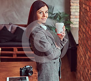 Young girl photographer dressed in a gray elegant jacket cup holding of takeaway coffee while leaning on a table in a