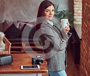 Young girl photographer dressed in a gray elegant jacket cup holding of takeaway coffee while leaning on a table in a