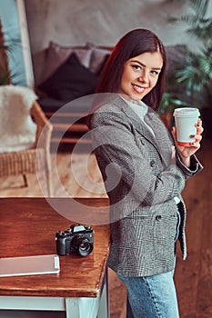 Young girl photographer dressed in a gray elegant jacket cup holding of takeaway coffee while leaning on a table in a
