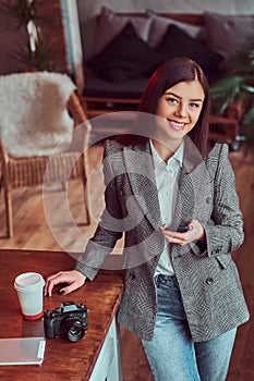 Young girl photographer dressed in a gray elegant jacket holding smartphone while leaning on a table in a room with loft