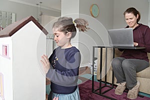 Young girl playing with doll house while her mother working on a laptop at home