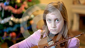 Young girl playing violin in front of christmas tree. Blonde young female musician violinist plays violin with Christmas