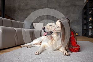 young girl plays with dog breed golden retriever at home on floor, woman with pet together lies on mat near the sofa