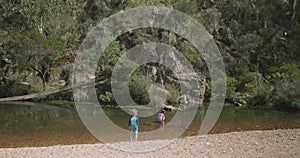 Young girl plays in a shallow stony pebbly river as boy walks down riverbank