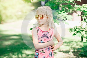 Young girl posing in fancy pink pentagonal shaped sunglasses outdoors. Cute serious pensive stylish Caucasian child with long