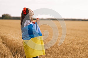 Young girl prays in the wheat field wearing Ukrainian national embroidered shirt with flower red wreath and holds flag at sunset y