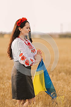 Young girl prays in the wheat field wearing Ukrainian national embroidered shirt with flower red wreath and holds flag at sunset y