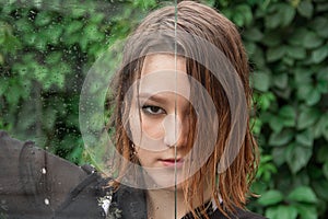 A young girl with predatory look and wet hair is looking at the camera. Wet glass with drops of water divides her face in half.