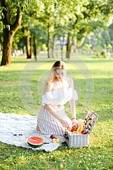 Young girl preparing forpicnic in park, sitting on plaid near box and waterlemon.