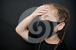 Young girl in protective sterile medical mask on her face has heat temperature, hold head with hand on a black background. Feeling