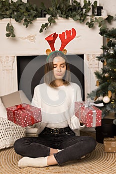 A young girl with puffy lips sits and holds two red Christmas gifts near the white fireplace