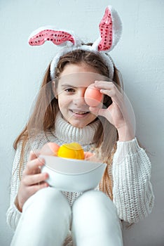 Young girl in rabbit ears holding easter egg with bowl of colorful eggs on white background