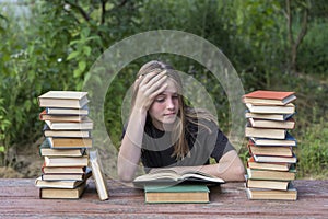 Young girl reading a book in the garden at a wooden table with a stack of books