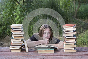 Young girl reading a book in the garden at a wooden table with a stack of books