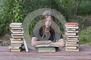 Young girl reading a book in the garden at a wooden table with a stack of books