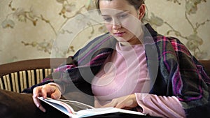 A young girl is reading a book sitting on the sofa in the living room. A beautiful girl in a pink sweatshirt is wrapped