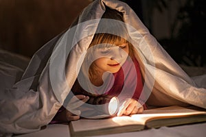 A young girl reading a book under the covers with a flashlight