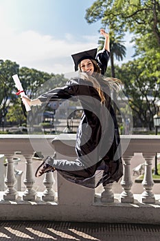 Young girl recently graduated, dressed in cap and gown, with her degree in her hand, jumping, on the university campus.
