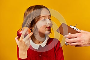 Young Girl in Red Choosing an Apple Over Chocolate Against a Yellow Background