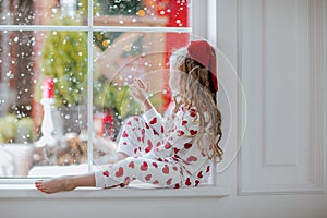 Young girl with red Christmas bow in white pyjamas posing on the window