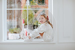 Young girl with red Christmas bow in white pyjamas posing on the window