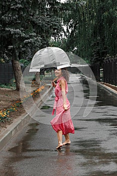 Young girl in a red dress with a transparent umbrella dancing in the rain standing in a puddle