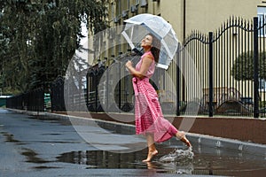 Young girl in a red dress with a transparent umbrella dancing in the rain standing in a puddle