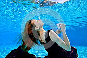 A young girl with red hair in a beautiful dress is swimming underwater in the pool on a blue background.