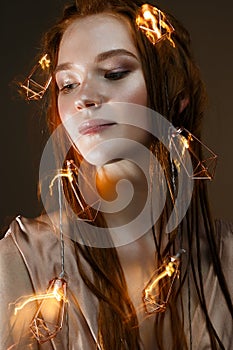 A young girl with red hair and with a luminous garland. Beautiful model with shining bulbs and lights. Gentle makeup.