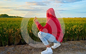 Young girl with red jacket and hood, squatting and thumb up in a rice field at sunset time. Concept like, approval, ok