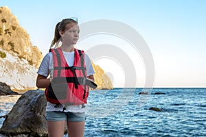 Young girl in red life jacket stands on beach with rock and sea. Female lifeguard on duty of coast. Water safety concept