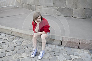 Young girl in a red wool sweater and jeans shorts is sitting on the steps