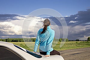 A young girl resting on the trunk of a car against the backdrop of an incipient thunderstorm, during a long journey on a suburban