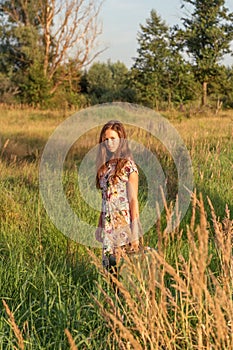 A young girl in a retro vintage dress with flowing hair is standing in a tall grass in a sunlight afterglow with a suitcase