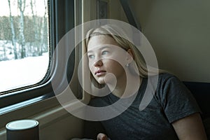 A young girl rides in a train compartment and looks out the window. Sad pretty blonde girl in a gray T-shirt. Close-up. Travel and