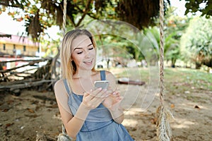 Young girl riding swing and using smatrphone, sand and tree in background.