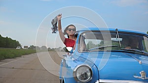 Young girl with scarf in hand leaning out of vintage car window and enjoying trip. Woman looks out of moving retro car