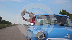 Young girl with scarf in her hands leaning out of window of vintage car and enjoying ride. Woman looks out from moving