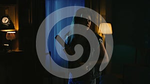 Young girl with scaried face expression standing in the dark living room, looking around, holding baseball bat.