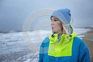 A young girl at the sea on a sunny day in winter. A pretty blonde in a blue hat and a bright sports jacket. Active recreation and