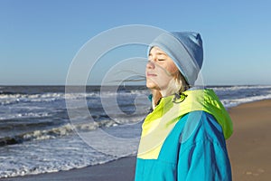 A young girl at the sea on a sunny day in winter. A pretty blonde in a blue hat and a bright sports jacket. Active recreation and