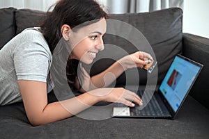 Young girl shopping online with credit card on laptop while lying on sofa