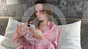 A young girl after a shower in the evening dries her hair sits on a bed in a hotel plays on a mobile phone uses the