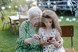 Young girl showing something on smartphone to elderly grandmother at garden party. Love and closeness between