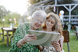 Young girl showing someting on tablet to elderly grandmother at garden party. Love and closeness between grandparent and