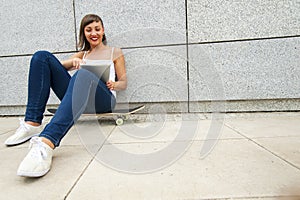 Young girl siting on skateboard in the cty with tablet.