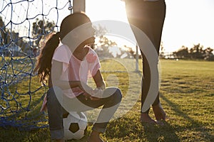 Young girl sits on ball next to her mum on a football pitch