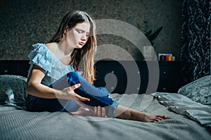 Young girl sits on a bed in her room, holds her injured leg with her hand